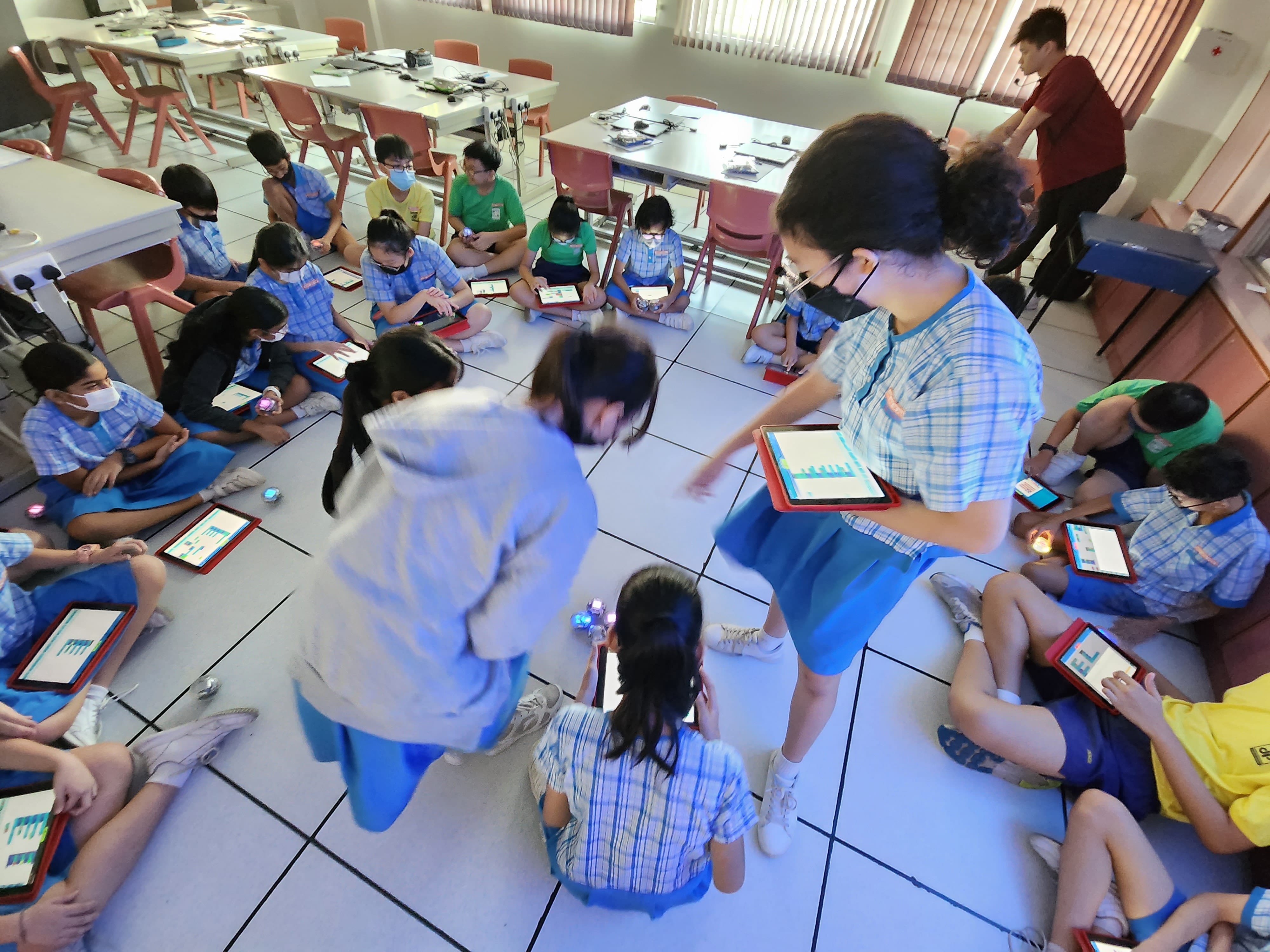 Students in blue uniforms and masks hold tablets, standing near small blue robotic balls on a tiled classroom floor.